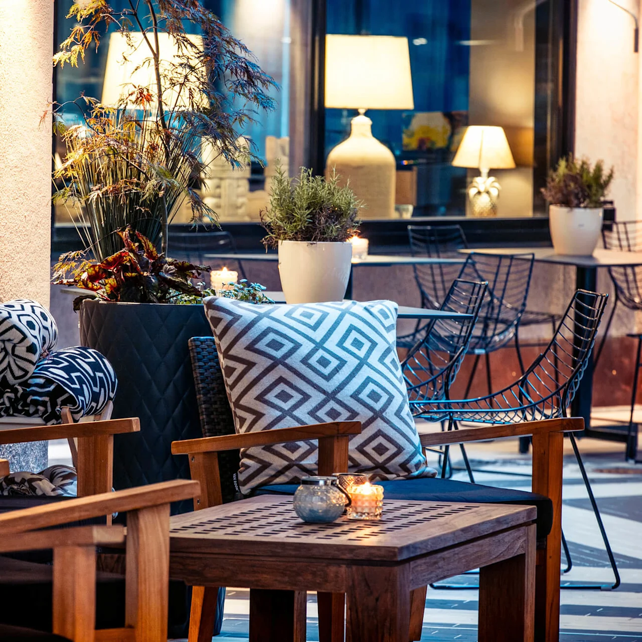 Trestoler med mønstrede puter sitter på en terrasse, omgitt av potteplanter og lys. Nærliggende bord og moderne stoler er arrangert utendørs i varm belysning.