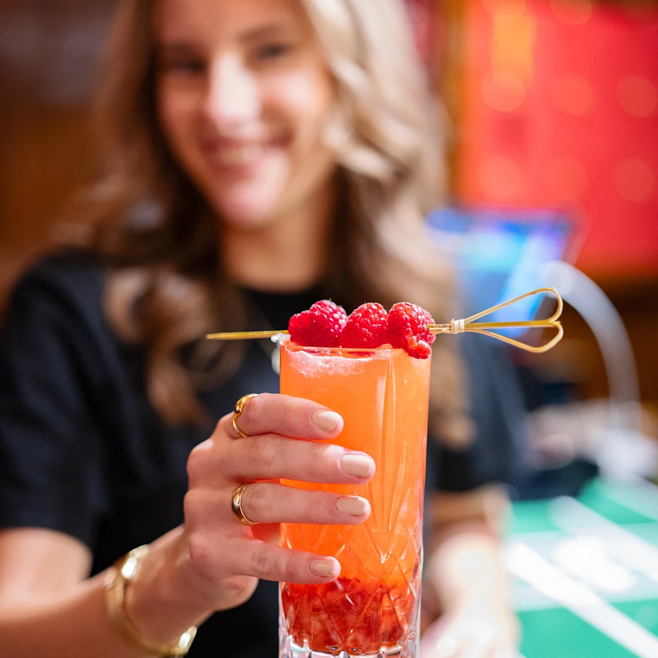 En person håller en livfull orange cocktail garnerad med hallon i ett högt glas, mot en livlig barmiljö med suddig bakgrund.