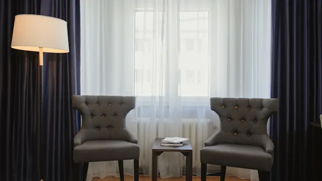 Två grå fåtöljer vänder sig mot ett litet bord med böcker, placerade i ett rum med en golvlampa och stort gardinklädd fönster, som skapar ett mysigt sittområde.