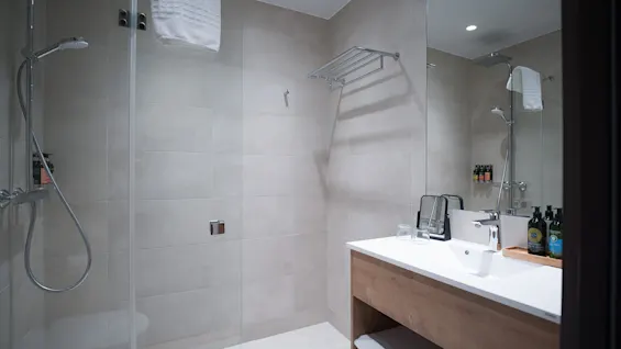 Shower with towel hanging, white sink with toiletries and mirror, in a modern, tiled bathroom context. Towels are neatly arranged in a shelf below the sink.