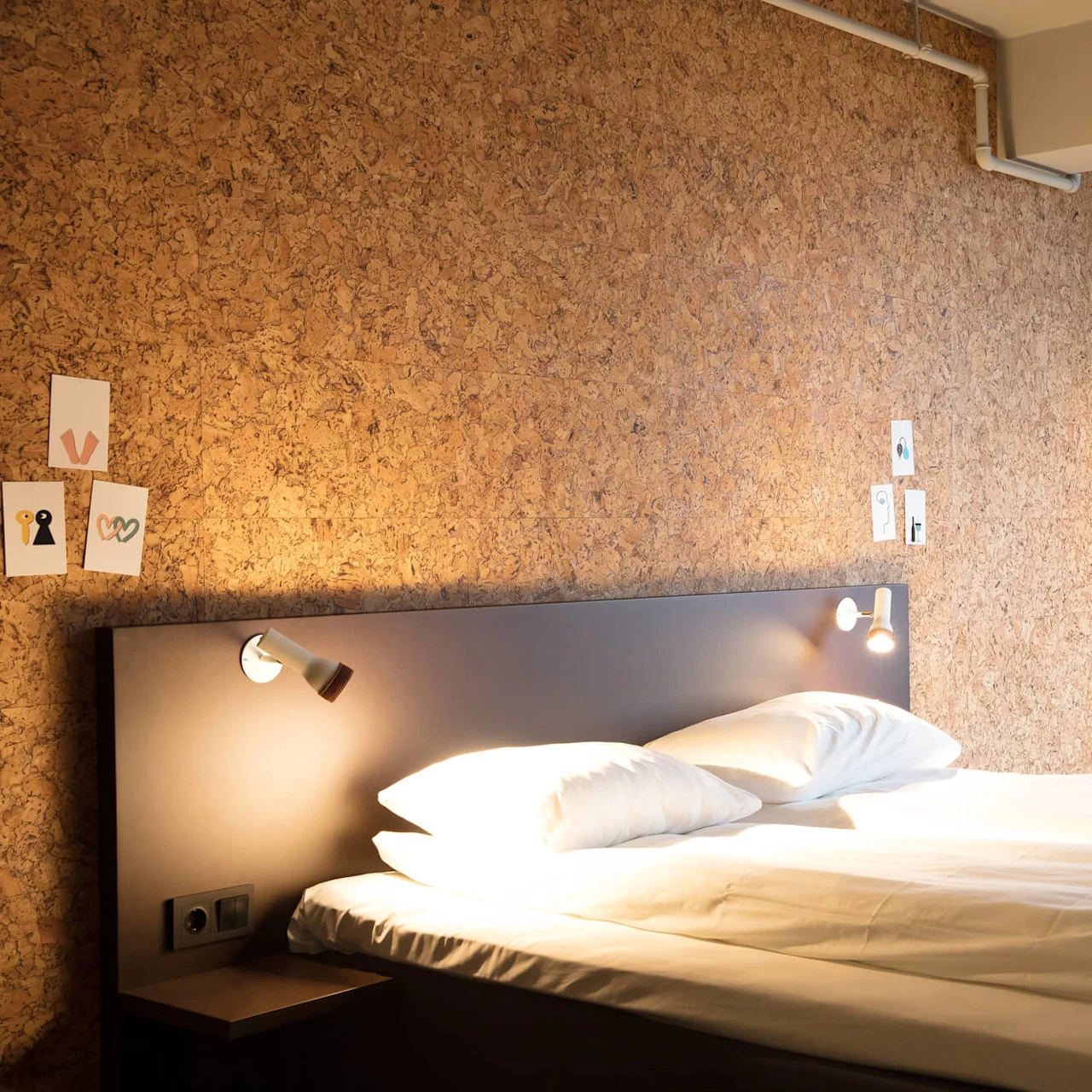 Bed with white sheets under two wall-mounted lights in a room. Decor includes minimalist art on a corkboard wall and a beige chair near a closed curtain.