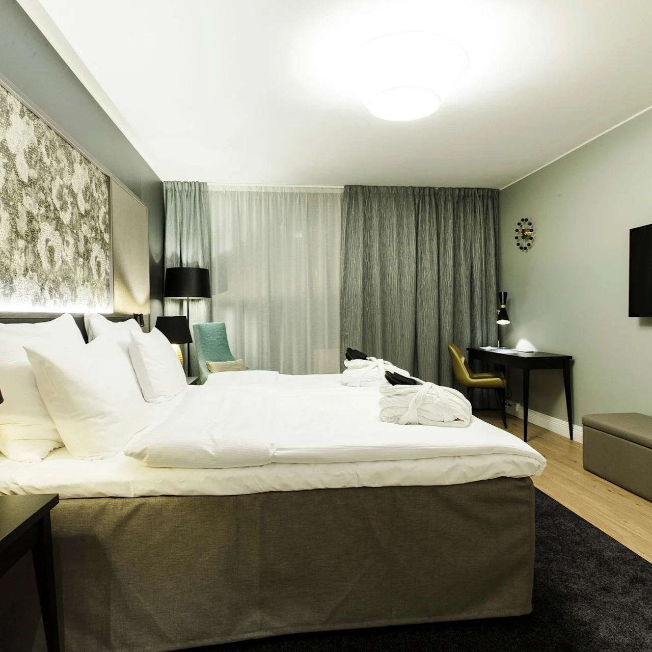 A neatly arranged hotel room with a large bed beside bedside lamps, facing a wall-mounted TV. A desk and chair are positioned near the window with draped curtains.