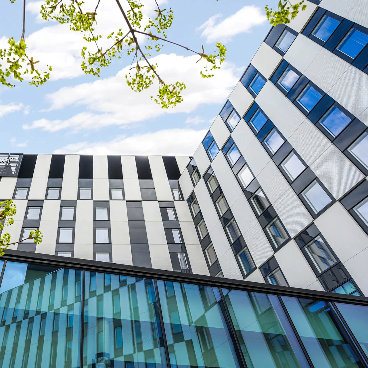 Building with geometric patterns stands under a blue sky. Green branches frame the view, creating a contrast with the architecture. The top displays "CLARION HOTEL & CONGRESS."
