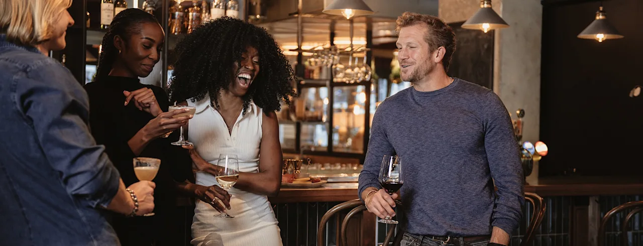 En gruppe venner nyder drinks og samtale på en livlig bar, med en person der griner glad.