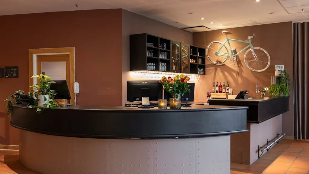 A stylish hotel reception and bar area with a curved counter, monitors, flowers, and a bicycle mounted on a brown wall.