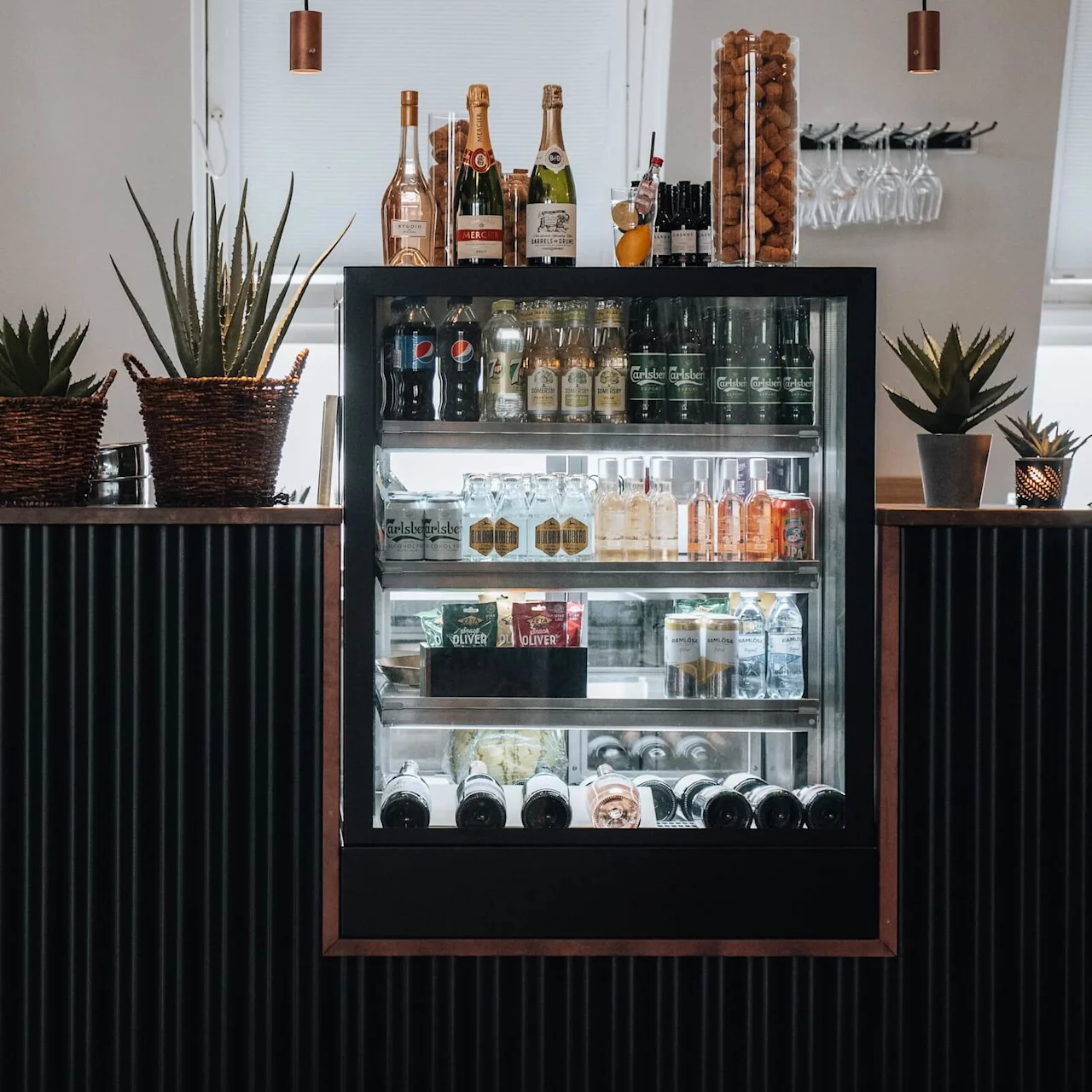 A black-framed refrigerator displays various beverages and snacks in a bar setting. Bottles and plants decorate the counter. Two menu signs are visible: "LASSE LUHAN DEN SAMLING DU ALDRIG FÅR" and "SELMA'S BAR MENU."