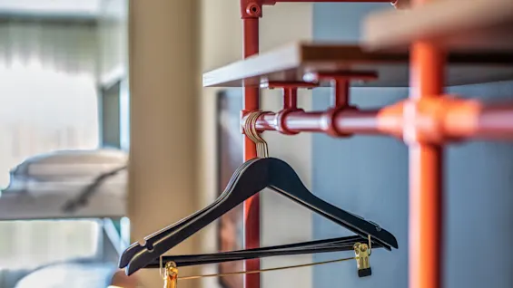 A black clothes hanger is suspended from a red industrial-style pipe rack in a modern room. Soft lighting filters through a window, and a bunk bed is partially visible.
