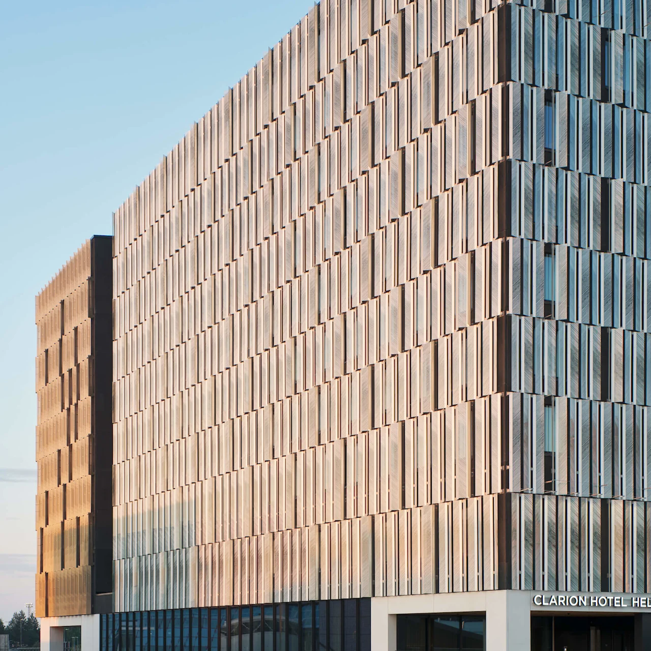 A modern building with a patterned facade reflects sunlight. "CLARION HOTEL HELSINKI AIRPORT" is written above the entrance. It's located in a quiet, mostly empty parking area.