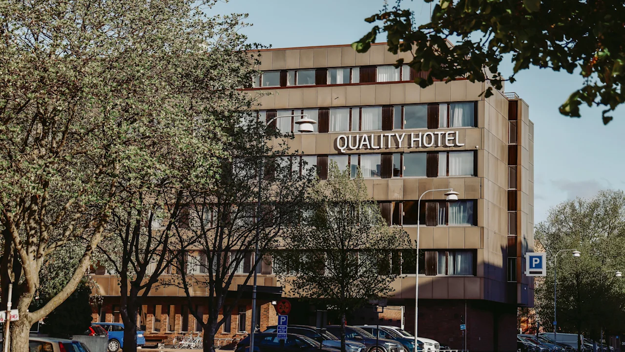 Quality Hotel sign on a modern, multi-story building surrounded by trees and parked cars. The trees are partly obscuring the view of the hotel, creating a natural urban setting.