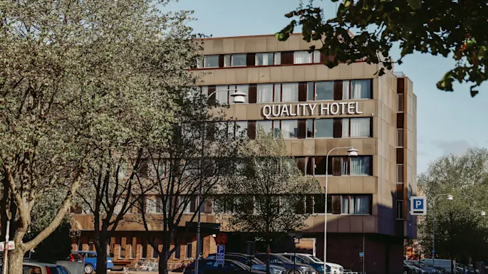 Quality Hotel-skilt på en moderne, fleretasjes bygning omgitt av trær og parkerte biler. Trærne skygger delvis for utsikten til hotellet, og skaper en naturlig urban setting.