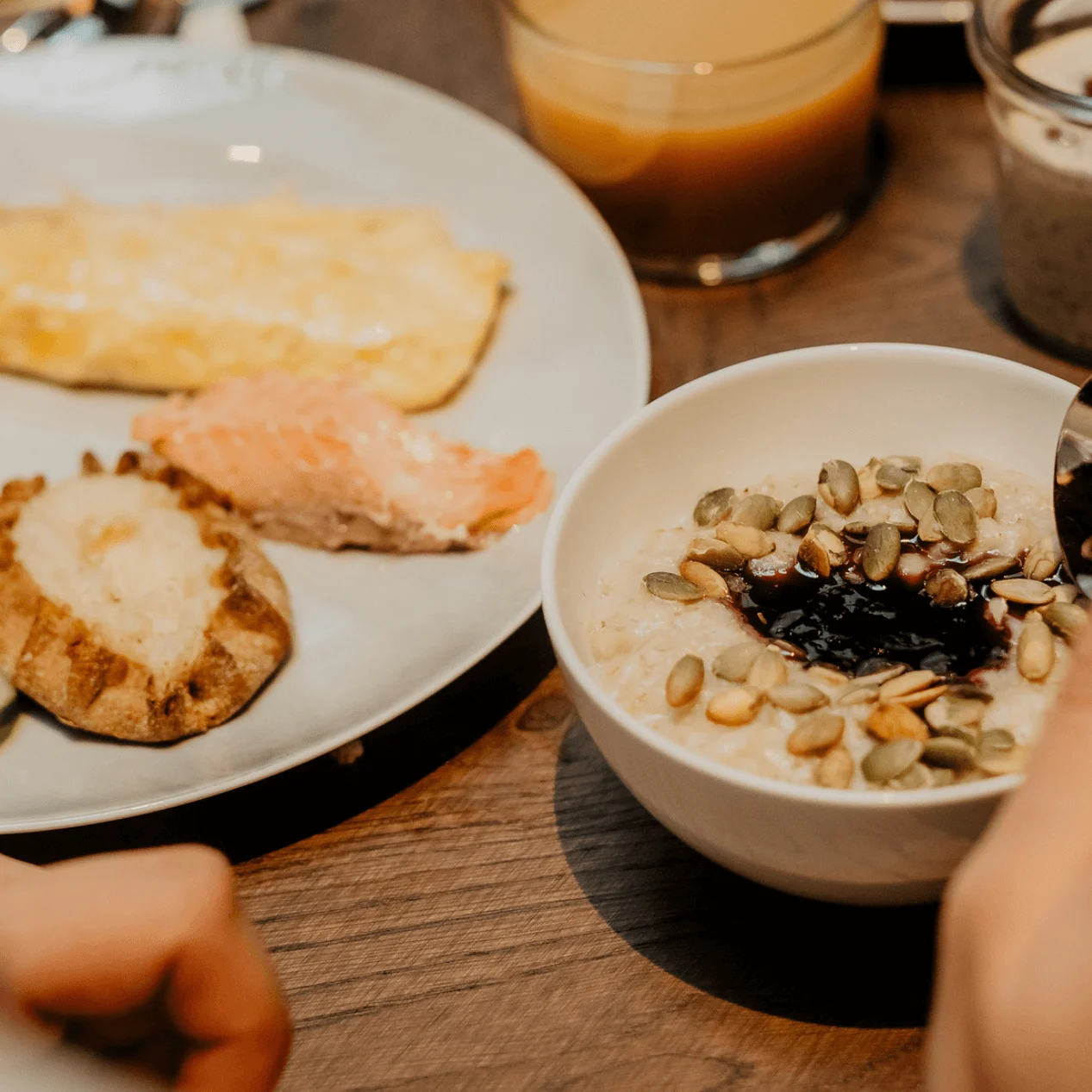 En skål med grød toppet med frø, omgivet af en hånd der holder en ske. Ved siden af er en tallerken med grøntsager, laks, omelet og kartoffel. Et glas juice og yoghurt kan også ses.