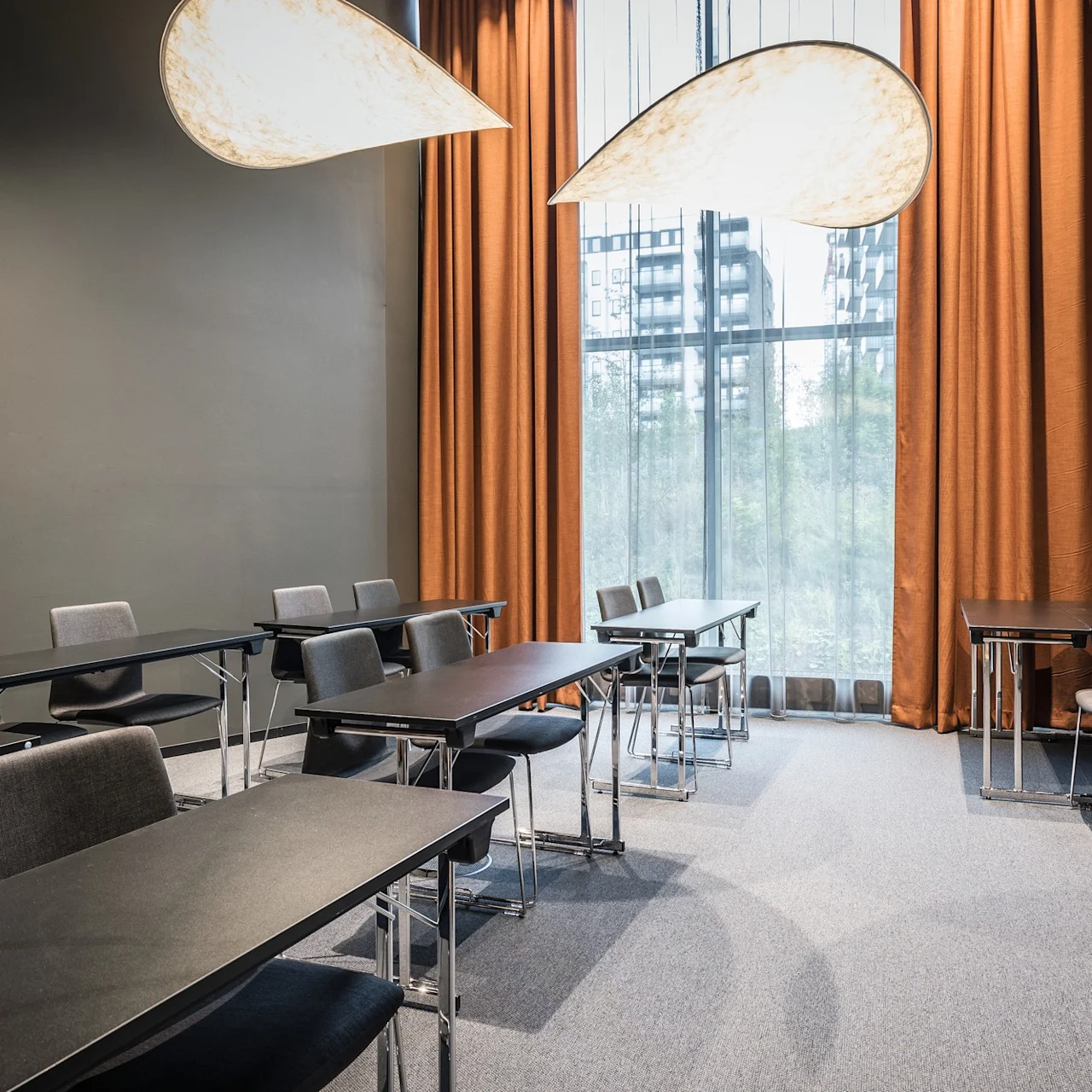 Klassrumsuppställning med rader av svarta bord och grå stolar, under stiliserade lövformade lampor. Stora fönster med orange gardiner med utsikt över moderna byggnader.