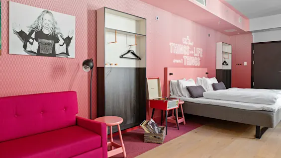Et levende hotellrom med rosa sofa, seng, åpen garderobe og veggkunst. Hvit neontekst: "The best THINGS in LIFE aren't THINGS." En platespiller står i nærheten.