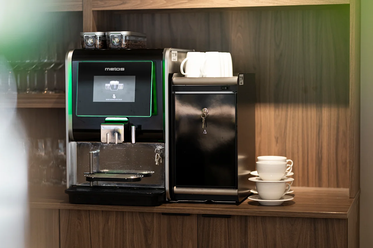 A modern coffee machine on a wooden counter, with coffee beans and stacked white cups ready for use. The screen shows 3 cups.