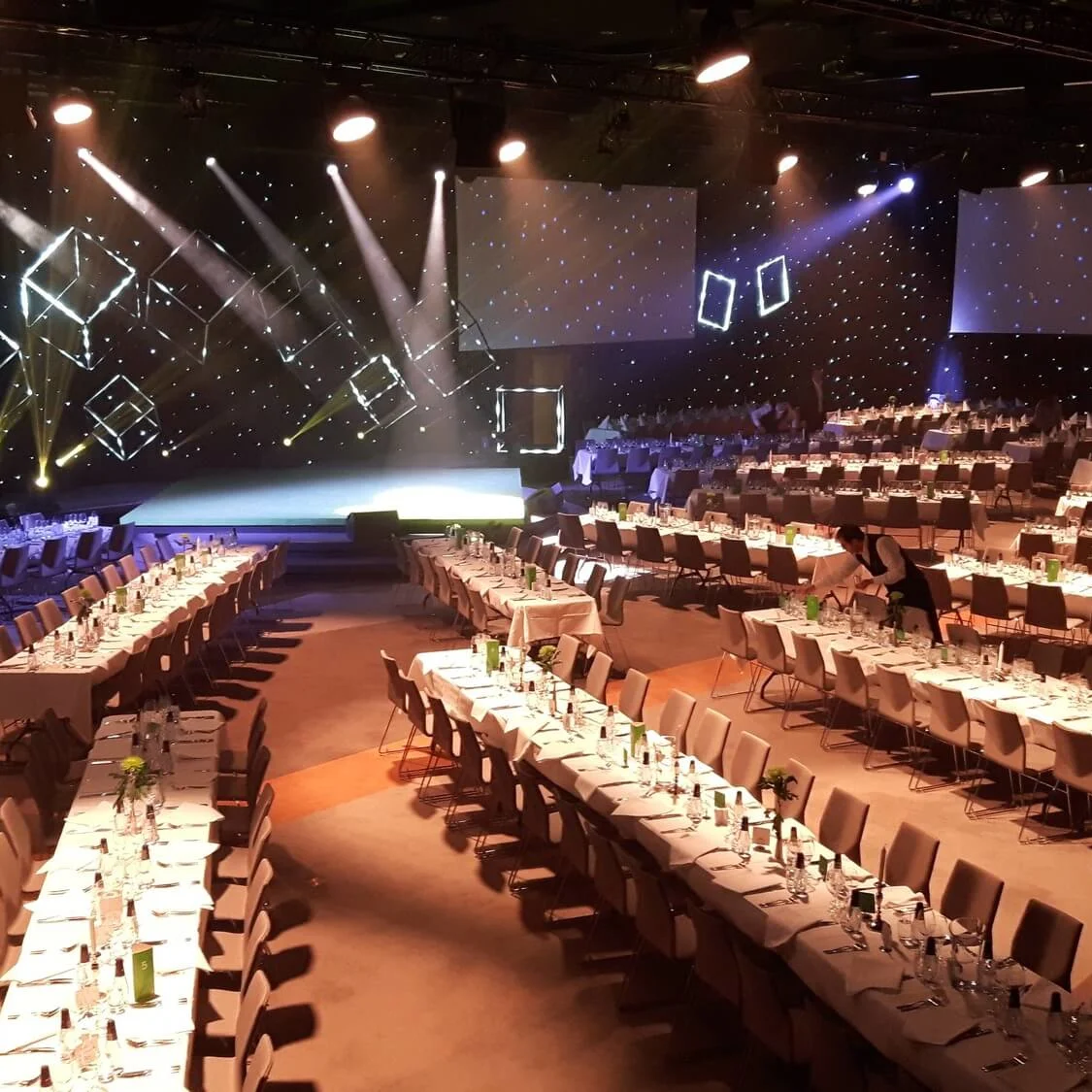 Tables are arranged in rows, elegantly set with glassware and cutlery, in a dimly lit banquet hall. Staff prepare for an event under decorative lighting and geometric designs.