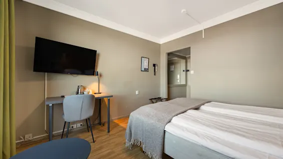 Lyst hotellrom med en dobbeltseng, et lite skrivebord med stol og en veggmontert TV.
