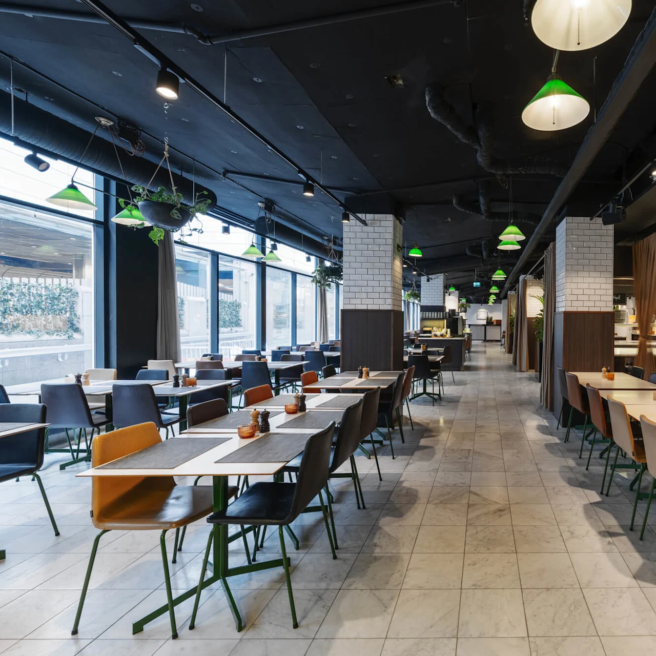 A restaurant interior features empty tables and mixed chairs, under green pendant lights, with a tiled floor and large windows. Potted plants hang from the ceiling.