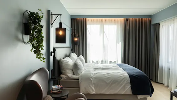 Et koselig hotellrom med en komfortabel seng, en stilig lenestol og et stort vindu med tynne og mørkleggingsgardiner.