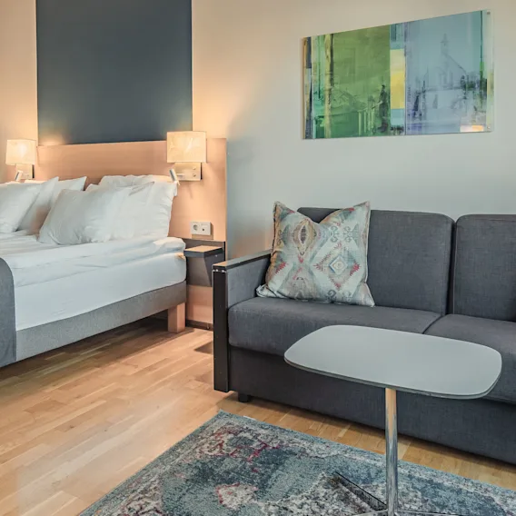 En säng och grå soffa med mönstrade kuddar är placerade i ett modernt sovrum. Trägolv och en matta ger värme, medan väggkonst och lampor skapar en mysig atmosfär.