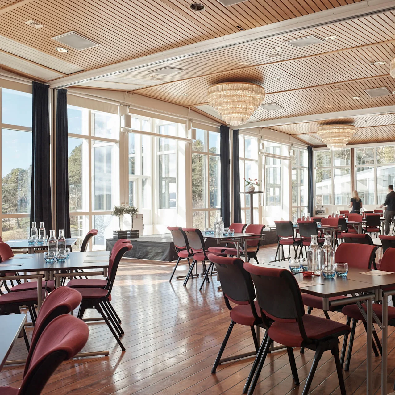 Bright room with red chairs and tables set for an event. People move in the background; large windows offer an outdoor view. Ceiling features chandeliers, a fruit bowl on the table.