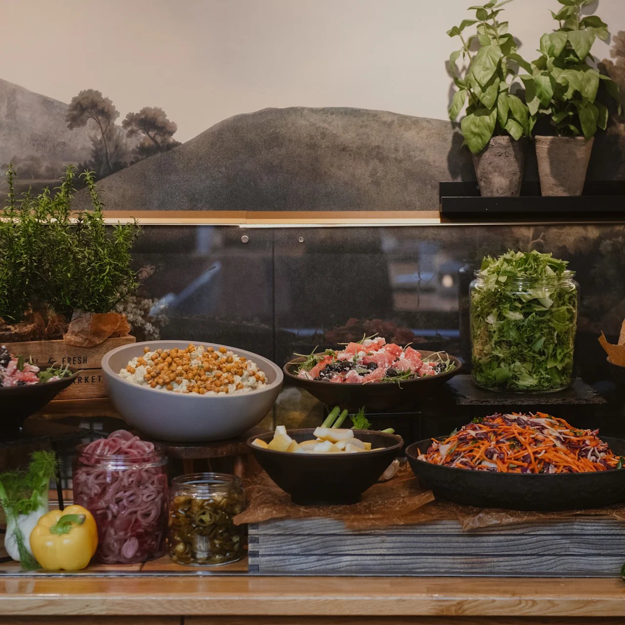 En række friske salater og grøntsager vises i skåle på et træbord. Urter og citroner er arrangeret ovenfor, mod et vægmaleri af et landligt landskab.