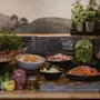 En mängd färska sallader och grönsaker visas i skålar på ett träbord. Örter och citroner är arrangerade ovanför, mot en muralmålning av ett lantligt landskap.