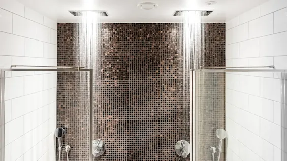 To takdusjer som strømmer vann i et moderne bad med hvite flislagte vegger og en sentral brun mosaikkaksent, omgitt av glassdører.