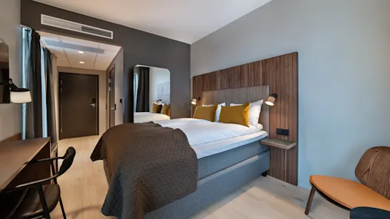 Et pent ryddet hotellrom med en oppredd seng, gule puter, tre-sengegavl og sengelamper. En pult og stol står ved siden av et vindu med mørke gardiner.