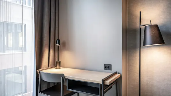 Ett skrivbord i trä med en svart stol och lampa placerad bredvid ett fönster med gardiner och en stående golvlampa i ett modernt, minimalistiskt rum.