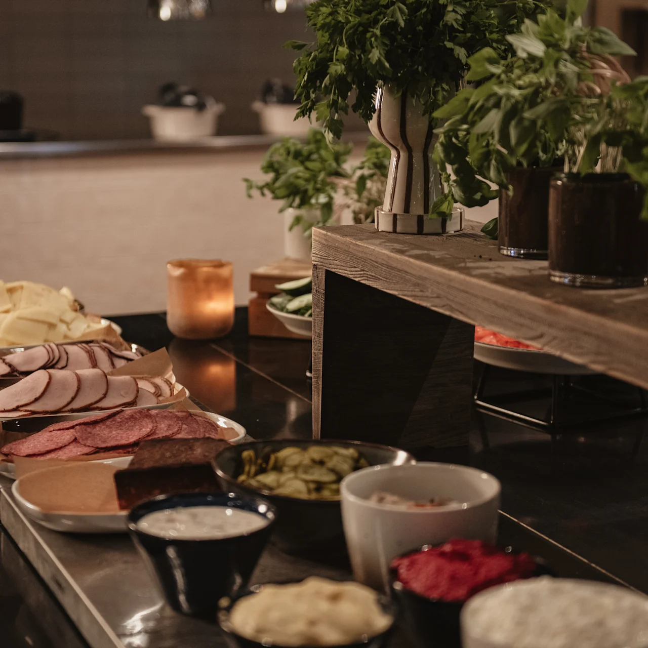 Various sliced meats and cheeses arranged on a wooden table, surrounded by small bowls of condiments; potted herbs accent the setting in a cozy, warmly lit kitchen.