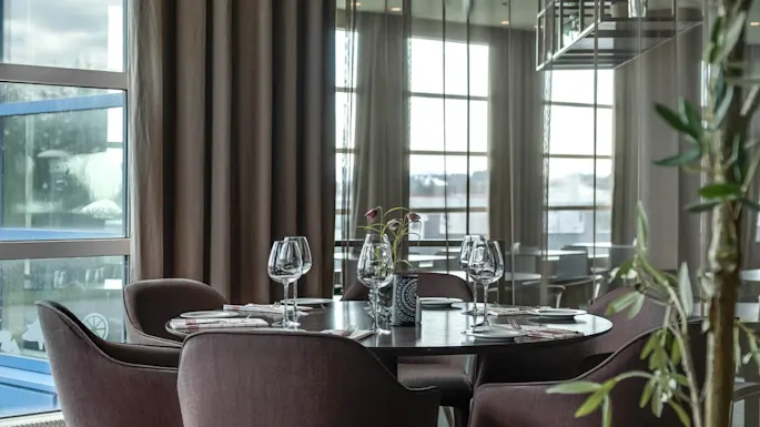 A round dining table set for four, with empty wine glasses and cutlery, in an elegant room with large windows and curtains.