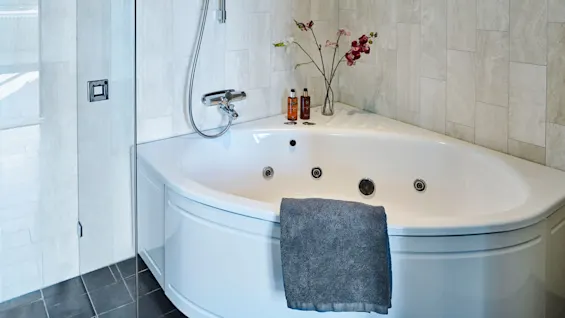 Et hvitt hjørnebadekar med jetstråler har et grått håndkle. I konteksten omgir elegante beige fliser, med blomster og toalettartikler på kanten, nær en glassdusjdør.