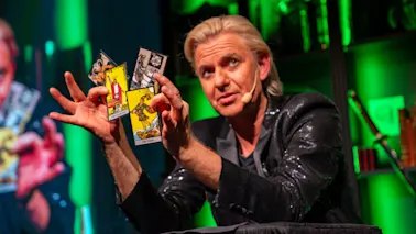 En person utför ett trolleritrick eller en tarotläsning, håller flera kort utspridda i handen på en grönbelyst scen.