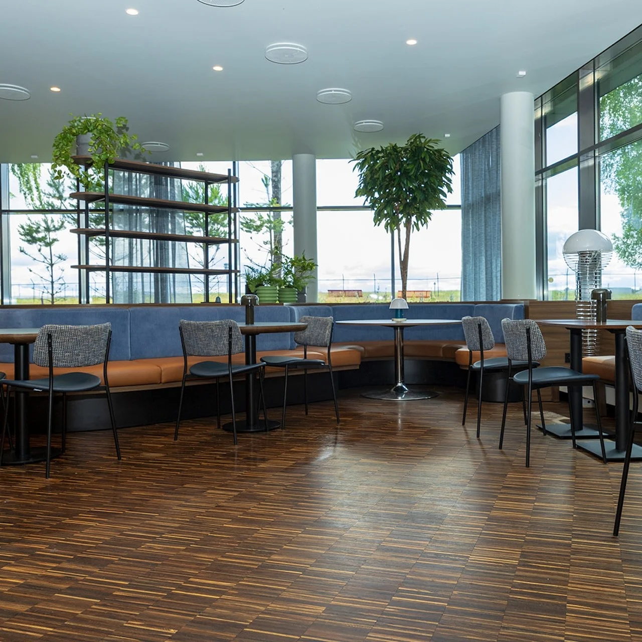 Modern café shows empty seating with round tables, cushioned chairs, and natural lighting through large windows. Indoor plants and wooden flooring create a fresh, inviting atmosphere.