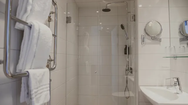 Duschområde med glasväggar innehåller duschhuvud och handtag. I närheten finns en handdukshängare med vita handdukar. Badrumsskiva med handfat, speglar och glas är bredvid. Vita kaklade väggar.