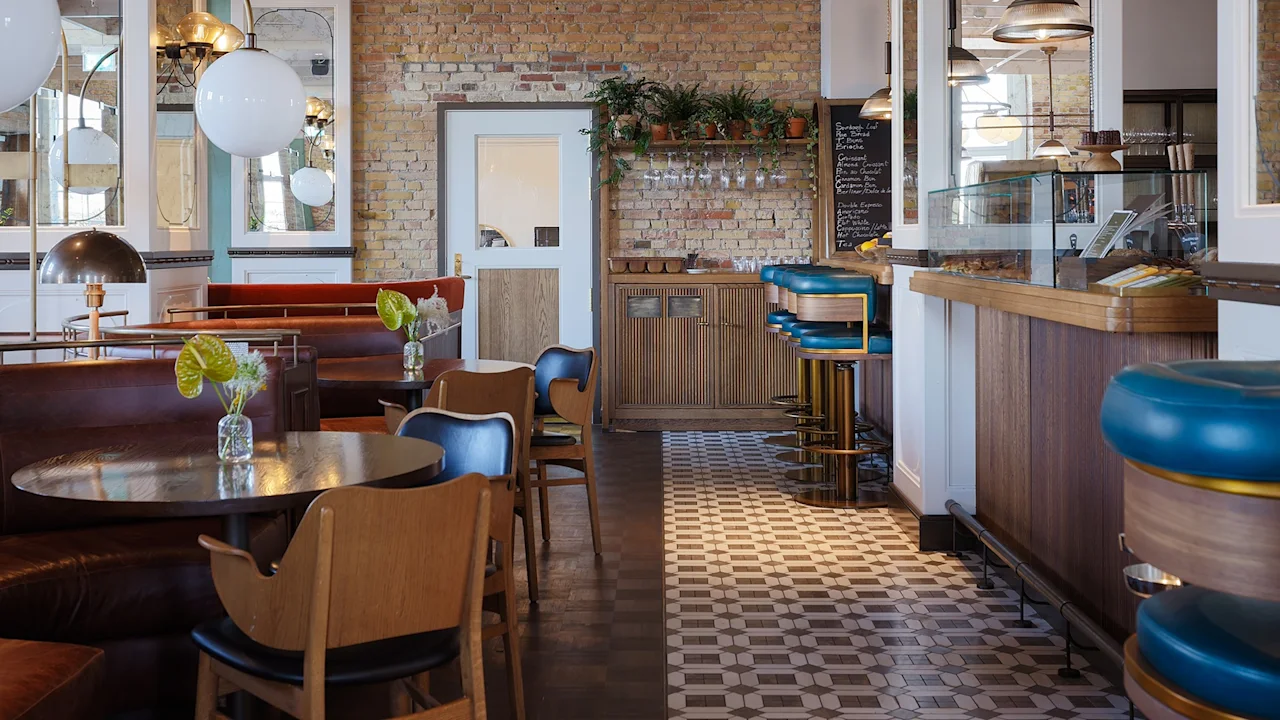 Et hyggeligt caféinteriør med træborde og stole med vaser af blomster. Barstole langs disken på et ternet flisegulv. Murstensvægge og store lamper skaber en varm atmosfære.
