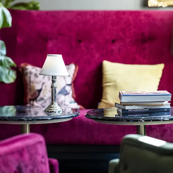 Et hyggeligt siddeområde med en magenta sofa, lænestole, små borde, en lampe og bøger, der indikerer et behageligt sted at slappe af.