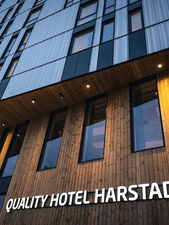 En hög byggnad med trä- och metallfasad, visar skylten "QUALITY HOTEL HARSTAD" upplyst. Sedd underifrån på kvällen, framhäver modern arkitektur.