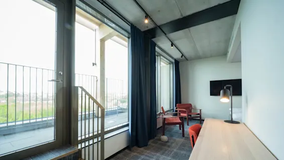 Moderni hotellihuone, jossa puupöytä ja lamppu, kaksi punaista tuolia suurten ikkunoiden vieressä, parvekkeelta näkymä vehreyteen.