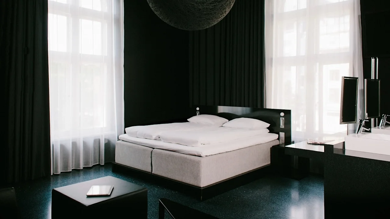 En väl bäddad säng mot en mörk vägg, med genomskinliga gardiner som mjukt filtrerar solljus i ett modernt sovrum.
