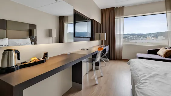 A hotel room features a desk with a kettle, tea packets, and a wall-mounted TV. A large window reveals a cityscape view. Text on pillow: "LOOK GREAT".