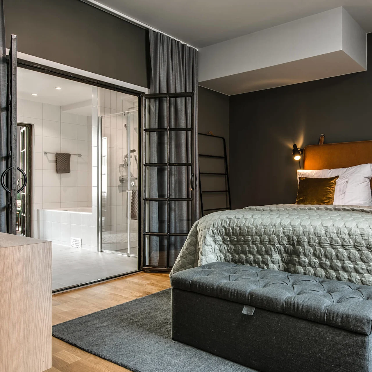 En seng med en tufted sengegavl står op ad en mørk væg, med grå og orange puder. Den vender mod et glasvægget bad i et moderne, varmt belyst værelse.