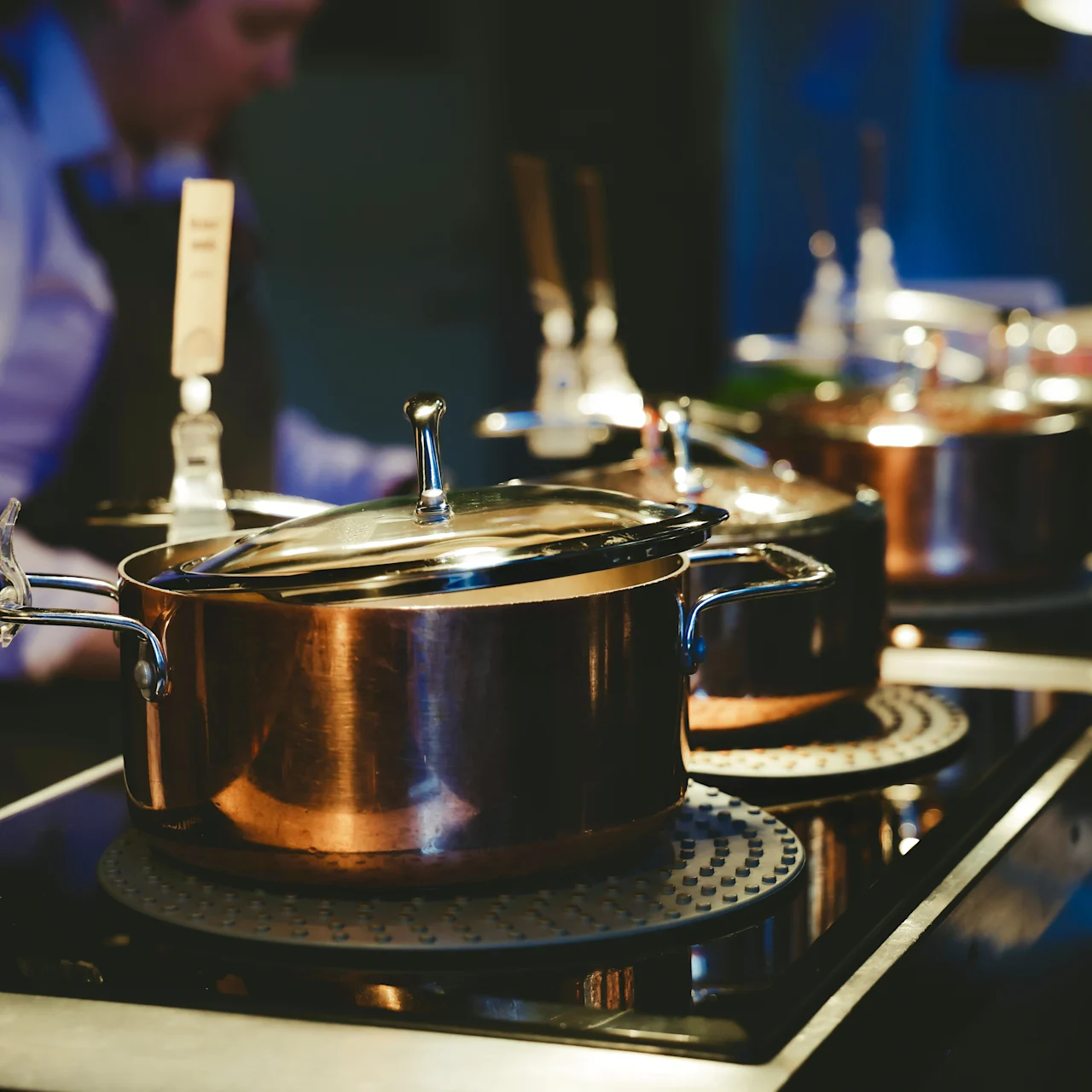 Copper pots with lids simmer on an electric stove in a dimly lit kitchen. A person in a white shirt stands nearby, preparing food.