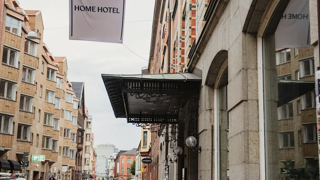 En travl europæisk gadescene med et HOME HOTEL-banner, historisk arkitektur og folk, der nyder en gåtur eller cykeltur.