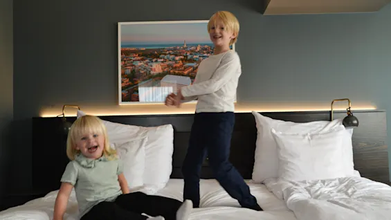 To børn hopper og sidder på en hvid hoteltseng, med et bybillede på væggen bag dem og blød stemningsbelysning.
