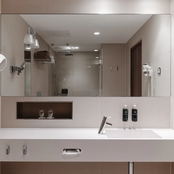 Ett modernt hotellbadrum med en stor spegel som reflekterar en dusch, en vit kommod med handfat, toalettartiklar och en hårtork.
