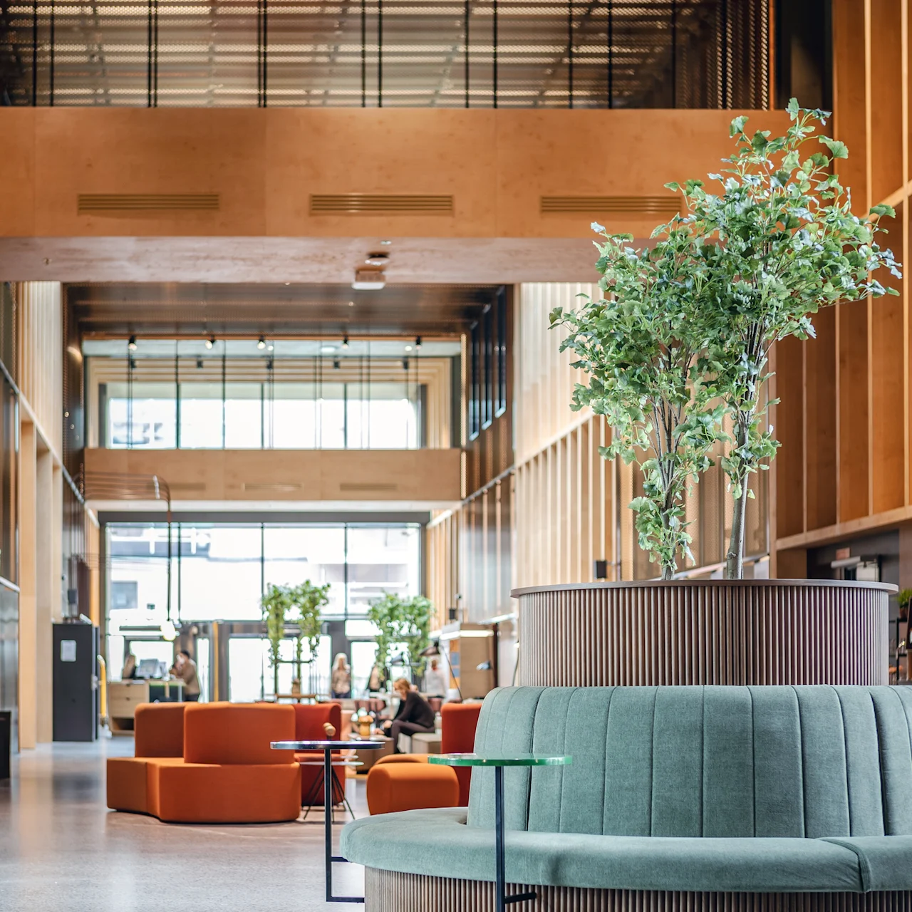 En cirkulär turkos bänk omger en plantering med en hög växt. Människor sitter i en modern, rymlig lobby med träväggar, glaselement och bekväma sittplatser i olika färger.