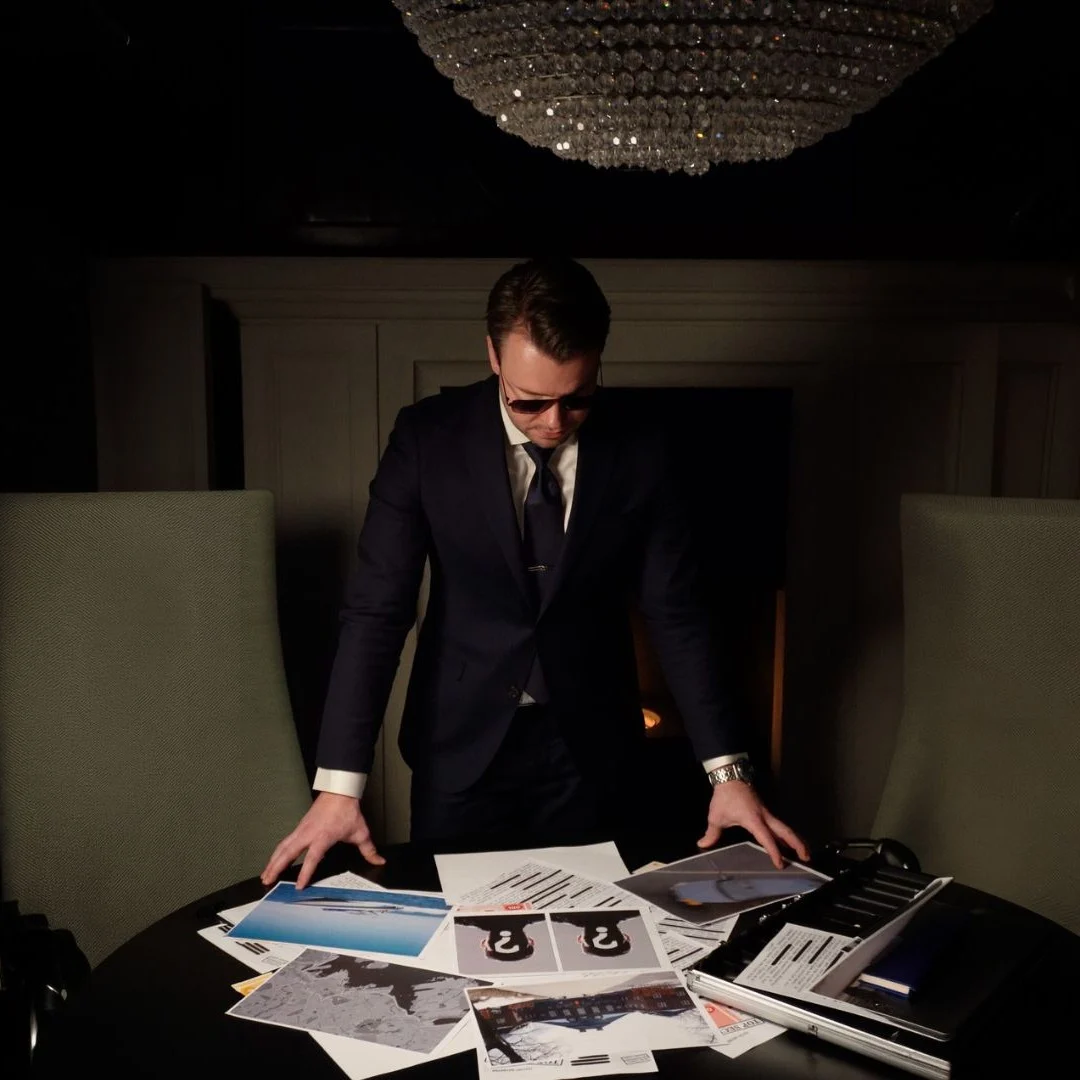 En person i dress og solbriller gransker nøye dokumenter spredt utover et bord i et dunkelt rom.