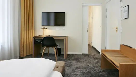 Et moderne hotelværelse med en seng, et skrivebord med stol, et vægmonteret TV og en træbænk. En døråbning fører til et badeværelse.