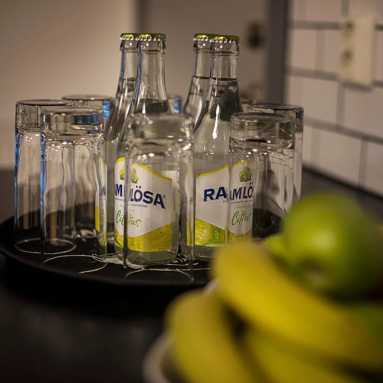 Empty glasses on a tray and citrus-flavored Ramlösa bottles on a kitchen counter, with a blurred fruit bowl in the foreground.